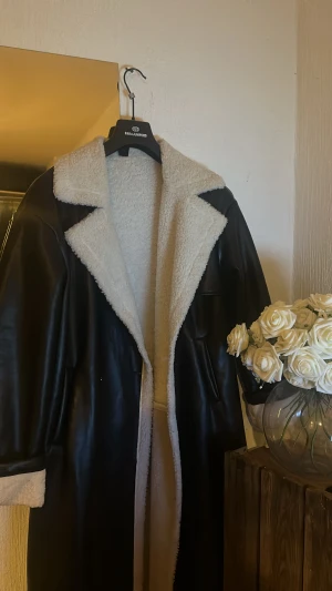 Svart teddyrock med vita detaljer - Säljer en svart lång teddyrock med vita fluffiga detaljer på krage, insida och ärmslut. Jackan har en oversized passform, dubbla knappar framtill och stora fickor. Utsidan är i skinnimitation och insidan är mjuk teddy. Perfekt för kalla dagar!