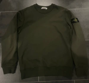 Mörkgrön Stone Island sweatshirt - Säljer en mörkgrön sweatshirt från Stone Island med klassisk rund hals och ikonisk patch på vänster ärm. Tröjan har ribbade muddar och är tillverkad i mjuk bomull, perfekt för en clean och stilren look.