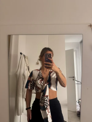 Mönstrad omlott blus med knyt - Säljer en croppad blus med korta ärmar och knyt framtill. Blusen har ett coolt mönster med inslag av leopard, kedjor och färger som vit, blå och brun. Perfekt för dig som gillar statement-plagg och vill sticka ut lite extra.