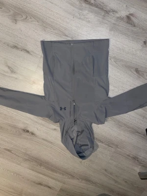 Grå vindjacka från Under Armour - Snygg grå vindjacka från Under Armour med huva och hel dragkedja framtill. Jackan har två sidofickor och diskret logga på bröstet. Tillverkad i lätt polyester som är perfekt för blåsiga dagar. Passformen är normal och ärmarna är långa.