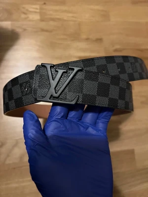 Svart Louis Vuitton bälte - Säljer ett stilrent svart bälte från Louis Vuitton med klassiskt rutigt mönster och en stor LV-logga i matt svart metall. Hör av er vid minsta fundering!