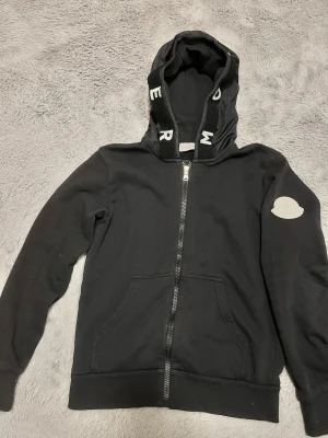 Svart Moncler hoodie med dragkedja - Svart hoodie från Moncler med vit logotyp på ärmen och coola bokstäver på huvan. Tröjan har hel dragkedja, fickor fram och mjukt fleecefoder på insidan. Huvan har detaljer i syntetmaterial och ribbade muddar vid ärmslut och nederkant.