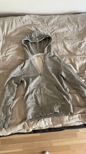 Grå hoodie från Champion med dragkedja - Grå hoodie från Champion med hel dragkedja och två stora fickor framtill. Mjuk insida och klassisk huva gör den perfekt för chill dagar. Diskret Champion-logga på bröstet och vid fickan. Passar dig som gillar enkel och clean stil.