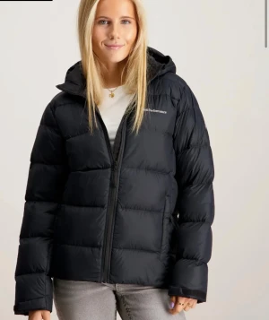 Svart pufferjacka från Peak Performance - Säljer en svart pufferjacka från Peak Performance med huva och dragkedja framtill. Jackan har en quiltad look och är tillverkad i syntetmaterial. Perfekt för kalla dagar och har en clean, modern vibe. Passformen är normal och jackan är långärmad.