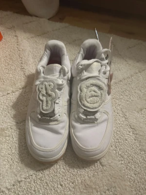 Nike Air Force 1 x Travis Scott – Vit (Cactus Jack Edition) - Nike Air Force 1 x Travis Scott – Vit (Cactus Jack Edition) 👟 Storlek: 42,5 ⚪ Färg: Vit ✨ Skick: 8/10 – använda men i bra skick 🔁 Detaljer: – Avtagbara Swoosh-märken i olika stilar (flera par ingår, ett silvrigt par saknas) – Avtagbar patch med Travis-logo över snörningen 📦 Ingår: Skorna + alla synliga tillbehör (på bild) 