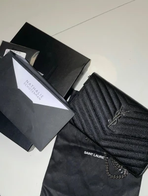 Svart kuvertväska Saint Laurent ÄKTA - Snygg svart kuvertväska från Saint Laurent med silverfärgad kedja och YSL-logga framtill. Väskan är quiltad i ett chevronmönster och har flera fack, inklusive dragkedja och kortplatser. Tillverkad i skinn med elegant struktur och magnetlås. Äkthetsbevis dustbag och kartong medföljer. Säljes i butik för 1430 euro. Motsvarar ca 16800 i svenska kronor. 