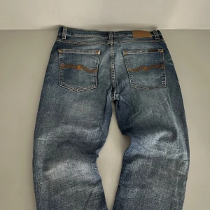 Blå jeans från Nudie Jeans Co - Säljer ett par klassiska blå jeans från Nudie Jeans Co med snygga slitningar och kontrastsömmar. Byxorna har fem fickor och en normal passform. Perfekta för dig som gillar en tidlös jeanslook.obs de finns ett litet hål på byxorna 