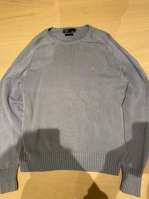 Ljusblå stickad tröja Polo Ralph Lauren - Snygg ljuvs blåa stickad  tröja från Polo Ralph Lauren i 100% bomull. Klassisk rund hals och ribbade muddar vid ärmslut och nederkant. På bröstet finns den ikoniska broderade loggan i gult. Perfekt för dig som gillar stilrena och tidlösa plagg. Den passar någon som bär L eller XL