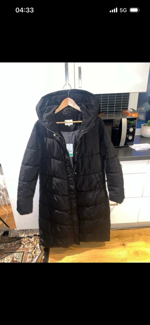 Svart lång pufferjacka från Aselle - Svart pufferjacka från Aselle i lång modell med huva och dragkedja framtill. Jackan har quiltad design och är tillverkad i syntetmaterial, perfekt för kalla dagar. Snygg och enkel stil som funkar till många outfits.