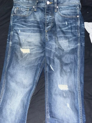 Blå slitna jeans från Jack & Jones - Säljer ett par blå jeans från Jack & Jones med snygga slitningar och lagade detaljer framtill. Klassisk femficksmodell med kontrastsömmar och läderpatch bak i midjan. Jeansen har en rak passform och är tillverkade i robust denim. W31 L34