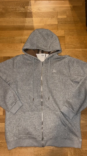 Grå hoodie från Burberry med dragkedja - Grå hoodie från Burberry med klassiskt rutigt foder i huvan och snyggt broderad logga på bröstet. Tröjan har hel dragkedja, snörning i huvan och två fickor framtill. Tillverkad i mjuk bomull för skön komfort. Perfekt för chill dagar. Köpt för 1500kr och säljs för 800kr för vill bli av med den snabbt pga den inte passar. Har aldrig använts. Skriv för fler frågor!