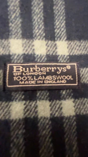 Rutig mörkblå halsduk från Burberrys - Snygg halsduk från Burberrys i klassiskt rutig design med mörkblå, grå och röda toner. Gjord av 100% lammull och har fransar i ändarna. Perfekt accessoar för att lyfta din stil under kyliga dagar.