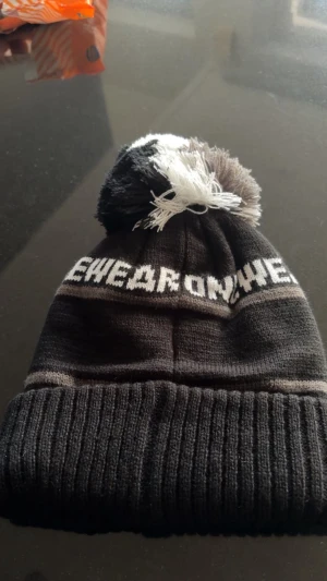 Svart Åre mössa med tofs och broderi - Snygg svart och grå stickad mössa från Onewear med stor tofs i svart, grått och vitt. Texten 'ÅRE' och bergmotiv broderat framtill ger en cool vibe. Perfekt för kalla dagar och vinterhäng.