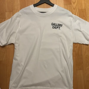 Vit Gallery Dept t-shirt  - Vit t-shirt från Gallery Dept. med svart tryck fram och stort tryck bak med texten 'GALLERY DEPT. HOLLYWOOD, CA.'. Klassisk rund hals och korta ärmar. Tillverkad i mjuk bomull för skön känsla. Perfekt för dig som gillar streetwear och enkel stil.