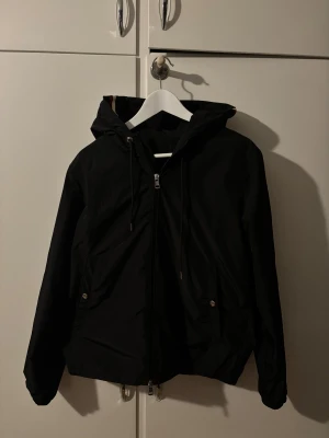 Svart vindjacka från Moncler - Svart vindjacka från Moncler med huva och dragkedja framtill. Jackan har snörning vid huvan, två fickor med knappstängning och Moncler-logga på vänster ärm. Snygga rödvita detaljer på huvan ger extra stilpoäng. Perfekt för dig som gillar clean och sportig look.