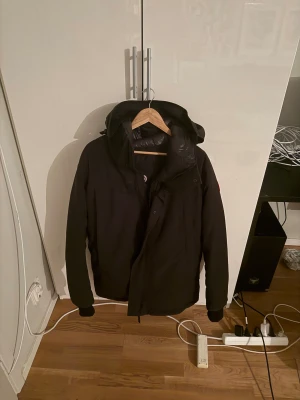 Svart vinterjacka från Canada Goose - Svart vinterjacka från Canada Goose med huva och klassisk logotyp på ärmen. Jackan har dragkedja och knappar framtill, ribbade muddar vid ärmslut och en stilren, mörk design. Perfekt för kalla dagar med sin tjocka och värmande fyllning.