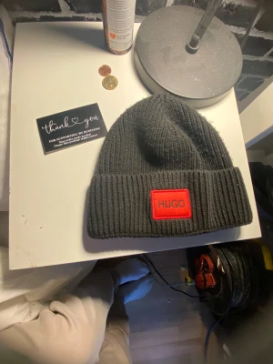 Hugo boss  - Snygg svart ribbstickad mössa från Hugo med en fet röd patch framtill där det står HUGO. Perfekt för dig som vill ha en clean och stilren look med lite edge. Materialet är mjukt och värmande, och mössan har en bred uppvikt kant.