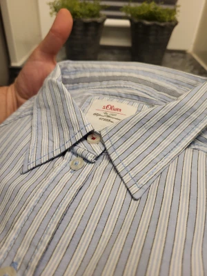 Randig skjorta från s.Oliver, stl 42 - Snygg skjorta från s.Oliver i ljusblått och vitt med tunna ränder. Klassisk krage, långa ärmar och knäppning framtill. Perfekt för dig som gillar stilrena och fräscha plagg. Skjortan är i mjuk bomull och har en clean look.