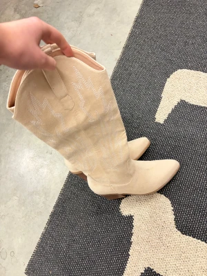 Beiga cowboy boots med spetsig tå - Snygga beiga cowboy boots i mocka-look med dekorativa sömmar och spetsig tå. Skorna har klassisk westernstil, perfekta för att lyfta vilken outfit som helst. Aldrig använda 