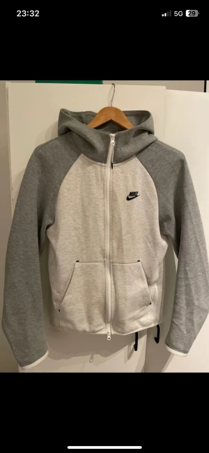 Grå och vit Nike hoodie med dragkedja - Nike hoodie i grått och vitt med hel dragkedja framtill, huva och två fickor. Snyggt broderad Nike-logga på bröstet. Skön passform och mjukt material, perfekt för chill eller träning. Långärmad och stilren design som funkar året runt.