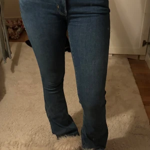 Blå bootcut jeans - Snygga blå jeans med bootcut passform och klassisk femficksdesign. Jeansen har låg midja, dragkedja och knapp framtill. De är lite slitna vid benslutet men bara på ena benet. Materialet är mjukt jeans-tyg som sitter bekvämt och följer kroppen.
