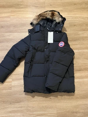 Svart dunjacka från Canada Goose - Svart dunjacka från Canada Goose med pälskantad huva och klassisk logga på ärmen. Jackan har quiltad design, dragkedja och knappar framtill. Perfekt för kalla dagar med sin tjocka och fluffiga look. Snygg och funktionell för dig som vill ha värme och stil.