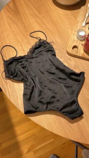 Svart satin body från Zara - Svart body från Zara i mjuk satin med tunna axelband och spetsdetaljer längs halsringningen. Snygg croppad passform som ger en cool vibe. Perfekt att styla med jeans eller kjol för en trendig look.