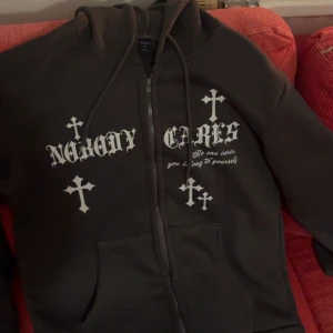 Svart hoodie med tryck från ROMWE - Svart hoodie från ROMWE i storlek S med vita kors och texten 'Nobody cares' tryckt på framsidan. Tröjan har huva med snörning, dragkedja och två fickor framtill. Perfekt för dig som gillar streetwear och edgy stil.