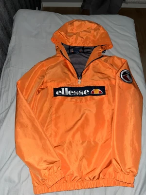 Orange vindjacka från Ellesse XS - Snygg orange vindjacka från Ellesse i storlek XS men passar S. Jackan har huva, half zip framtill med Ellesse-logga på dragkedjan, stor broderad Ellesse-logga på bröstet och patch på ärmen. Tillverkad i lätt polyester med meshfoder och elastiska muddar. 