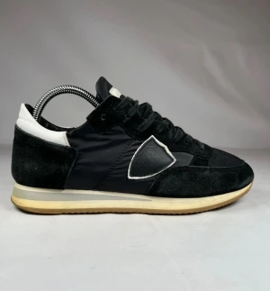 Svarta sneakers från Philippe Model - Svarta sneakers från Philippe Model med klassisk låg siluett. Skorna har en mix av mocka och textil, vit detalj på häl och plös samt den ikoniska skölden på sidan. Platt sula i beige och svart, snörning framtill och rund tå. Perfekt för dig som gillar stilrena och sportiga vibes.