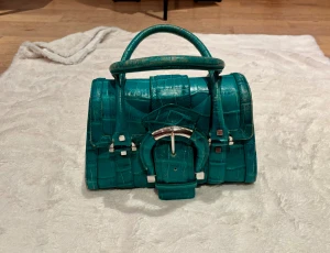 grön handväska med krokoprägling. Karen Millen - Cool klargrön handväska i croco-mönstrat skinn med silverfärgade metalldetaljer och spänne framtill. Väskan har två stadiga handtag och en unik, strukturerad form som sticker ut. Perfekt för dig som vill ha en statement-accessoar. Väskan är väldigt fräsch också inuti. Se bilder. Köpt på bilblioteksgstan i sth, nypris över 7000 sek