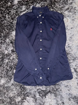 Mörkblå skjorta från Polo Ralph Lauren - Snygg mörkblå skjorta från Polo Ralph Lauren med klassisk krage och knappar framtill. Skjortan har lång ärm och den ikoniska röda broderade loggan på bröstet. Tillverkad i mjuk bomull som känns skön mot huden. Perfekt för dig som gillar stilrena och tidlösa plagg.