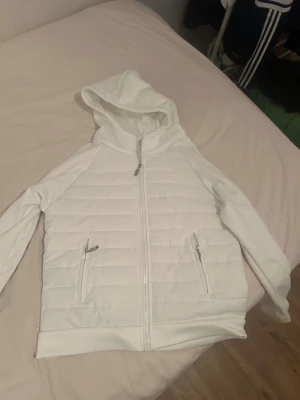 Vit cardigan - Säljer en vit cardigan med quiltad framsida och rygg, dragkedja hela vägen och två fickor med dragkedja. Jackan har huva och långa ärmar, perfekt för chilliga dagar. Materialet är polyester och syntet, vilket gör den både lätt och bekväm.