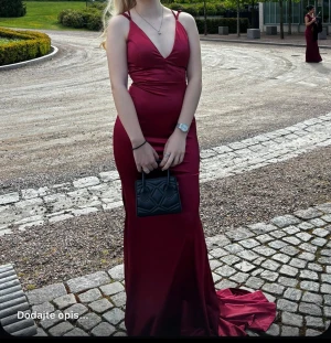 Vinröd långklänning med snörning - Säljer en vinröd långklänning i satin med djup v-ringning och smala axelband. Klänningen har snygg snörning och öppen rygg, samt figurnära passform och släp. Perfekt för bal eller festliga tillfällen.