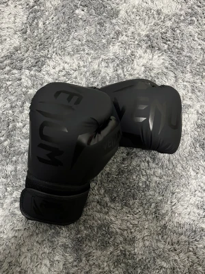Svarta boxningshandskar från Venum - Säljer ett par riktigt snygga svarta boxningshandskar från Venum. De har en matt finish med diskret Venum-logga i blankt svart på ovansidan och kardborrestängning vid handleden. Handskarna är vadderade och gjorda i syntetmaterial, perfekta för träning och sparring.