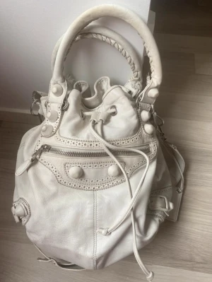  Balenciaga Pompom väska - Snygg vit axelväska från Balenciaga i mjukt skinn med flätade handtag, snörning upptill och dekorativa nitar. Väskan har dragkedja framtill, justerbar axelrem och ett fack med spegel. Superfint skick och rengjord med lädertvätt och insmord med läderbalsam❤️köpt för 6000 kr.