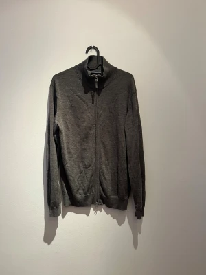 Grå kofta i merinoull från Hansen & Jacob - Snygg grå kofta från Hansen & Jacob i mjuk merinoull. Modellen har hel dragkedja framtill, ribbade muddar och hög krage. Elbow patches i matchande ton på ärmarna ger en cool detalj. Perfekt för lager-på-lager under höst och vinter.