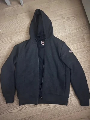 Mörkgrå hoodiejacka från Colmar - Snygg mörkgrå hoodiejacka från Colmar Originals med dragkedja och huva. Jackan har en stilren look med logga på ärmen och insida som håller vind. Perfekt för dig som gillar enkel och clean streetstil. Storleken är L Men jag skulle säga att den är bra för någon som använder storlek M. Den är som splitterny, har använt den bara ett par gånger på grund av att den blev för liten. 