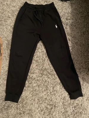 Svarta mjukisbyxor Polo Ralph Lauren 160 - Svarta mjukisbyxor från Polo Ralph Lauren med klassisk broderad logga i vitt på vänster ben. Byxorna har snörning i midjan, ribbade muddar vid bensluten och två sidofickor. Perfekta för chill eller träning, riktigt sköna och stilrena. Finns inga hål eller utslitna märken riktigt snygga och ser ut som att dom är nya