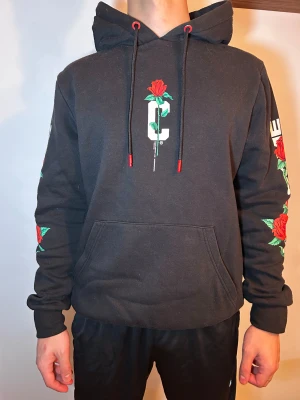 Svart hoodie med rosor och text - Svart hoodie från CROPP med stor vit text 'I NEED SOME NEW FRIENDS' på ryggen och röda rosor tryckta på både rygg, ärmar och bröst. Hoodien har huva med snörning och känguruficka framtill. Snygga röda detaljer på snörningen och coolt blommigt print.