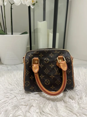 Monogram handväska - Säljer Monogram handväska i brunt skinn. Väskan har guldfärgad dragkedja och detaljer i läder med gul söm. Perfekt storlek för det viktigaste och riktigt trendig look. Med band och helt ny. 