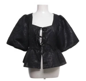 Svart glittrig blus med puffärm - Svart glittrig blus med stora puffärmar och snörning framtill. Peplum nedtill, Materialet har en skimrande finish som syns extra i ljuset. Perfekt nu inför nyår!🎇