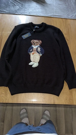 Svart stickad tröja Polo Bear - Svart stickad tröja från Polo Ralph Lauren med ikonisk Polo Bear broderad framtill. Rund hals, ribbade muddar och långärmad modell. Tröjan har en cool och klassisk vibe med detaljerad björn i blå och beige nyanser. Perfekt för dig som gillar street och preppy stil.