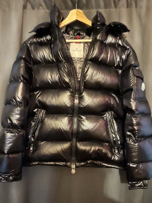 Svart Moncler pufferjacka med huva - Säljer en svart pufferjacka från Moncler med glansig finish och huva. Jackan har tvåvägsdragkedja med Moncler-logga, sidofickor med dragkedja och coolt serietryck på insidan. Perfekt för dig som vill ha en varm och snygg jacka med streetkänsla. Storlek 2 passar M 