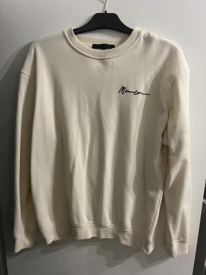 Beige sweatshirt från KENMACE - Snygg beige sweatshirt från KENMACE i storlek M. Tröjan har rund hals, ribbade muddar och ett stilrent svart tryck på bröstet. Perfekt för dig som gillar enkel och clean stil. Materialet är mjukt och skönt, troligtvis bomull eller polyester.