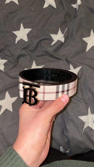 Burberry bälte - Säljer ett snyggt Burberry bälte med klassiskt rutig design i beige, svart och röd. Bältet har ett stort TB-spänne i guld och svart som verkligen sticker ut. Perfekt accessoar för att lyfta din outfit med lite lyxig vibe. 120cm, går att byta sida på bältet.