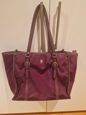 Mörklila/burgundy väska från U.S. Polo Assn. - Stilren mörklila totebag från U.S. Polo Assn. med guldiga detaljer och dubbla handtag. Väskan har dragkedja upptill, dekorativa spännen och logga framtill. Insidan är fodrad med mönstrat tyg och har flera fack för småsaker. Väska/shopper/totebag från US Poloa Assn i fin mörkröd/burgundy färg. Stor och rymlig och rymmer en laptop.  Dragkedjan går inte alltid igen och finns lite slitage (se bild), men i övrigt fint skick. 