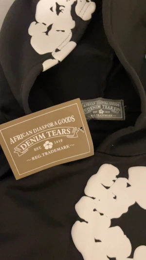 Svart hoodie från Denim Tears med tryck - Svart hoodie från Denim Tears med stora vita grafiska tryck i cirkelform över hela tröjan och huvan. Tröjan har huva och en klassisk känguruficka framtill. Materialet är mjukt och känns som bomull, perfekt för dig som gillar streetwear med unik design.