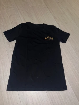 Svart t-shirt från Hugo Boss - Svart t-shirt från Hugo Boss med gul logotyp på bröstet. 