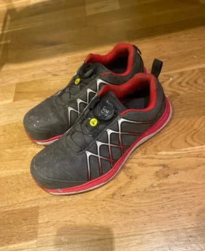  Stålhettaskor från Gesto med BOA-snörning - Säljer ett par svarta Stålhetta arbetsskor från Gesto med röda och vita detaljer. Skorna har BOA-snörning för enkel justering och en sportig look. Ovandelen är i syntetmaterial med mönstrade linjer och röd insida. Perfekta för dig som gillar stilrena och funktionella skor. Skorna är väldigt använda som man ser men går enkelt att rengöra med en tvätt. Storlek 38 